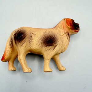 May include: A molded plastic dog figurine in a standing position. The figure is primarily cream-colored with dark brown patches on the back and sides, a dark brown face, and orange-toned ears and tail tip. The dog has a sculpted fur texture.