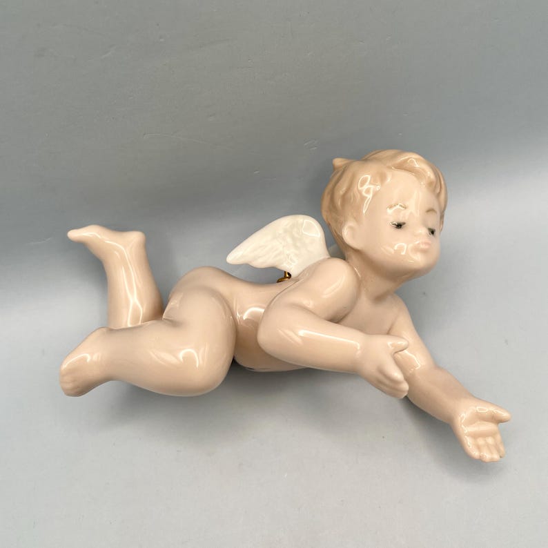 A porcelain figurine of a cherub, lying down with one arm outstretched. The cherub has a light skin tone, blonde hair, and small white wings. The figurine is a decorative item.