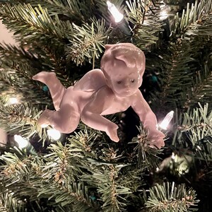 May include: A porcelain Christmas tree ornament in the shape of a baby. The baby is light pink and is crawling. The ornament is hanging on a green Christmas tree with white lights.