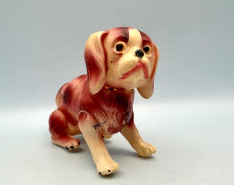 Op de afbeelding: Een vintage hondenbeeldje in een zittende positie. De speelgoedhond heeft een crèmekleurig gezicht en poten, met donkerrode en crèmekleurige vacht op zijn lichaam en oren. Het heeft een zwarte neus en ogen en een gele halsband. Het beeldje is gemaakt van hard plastic.