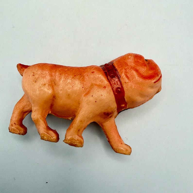 May include: A small, peach-colored plastic figurine of a bulldog standing in profile. The dog has a wrinkled face, a short tail, and a dark red collar with small, raised circular details around its neck. The surface has a smooth, molded texture.