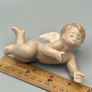 May include: A porcelain figurine of a cherub with closed eyes and small white wings. The cherub is lying down, with one arm extended. The figurine is a light pink color. A wooden ruler is in the foreground.