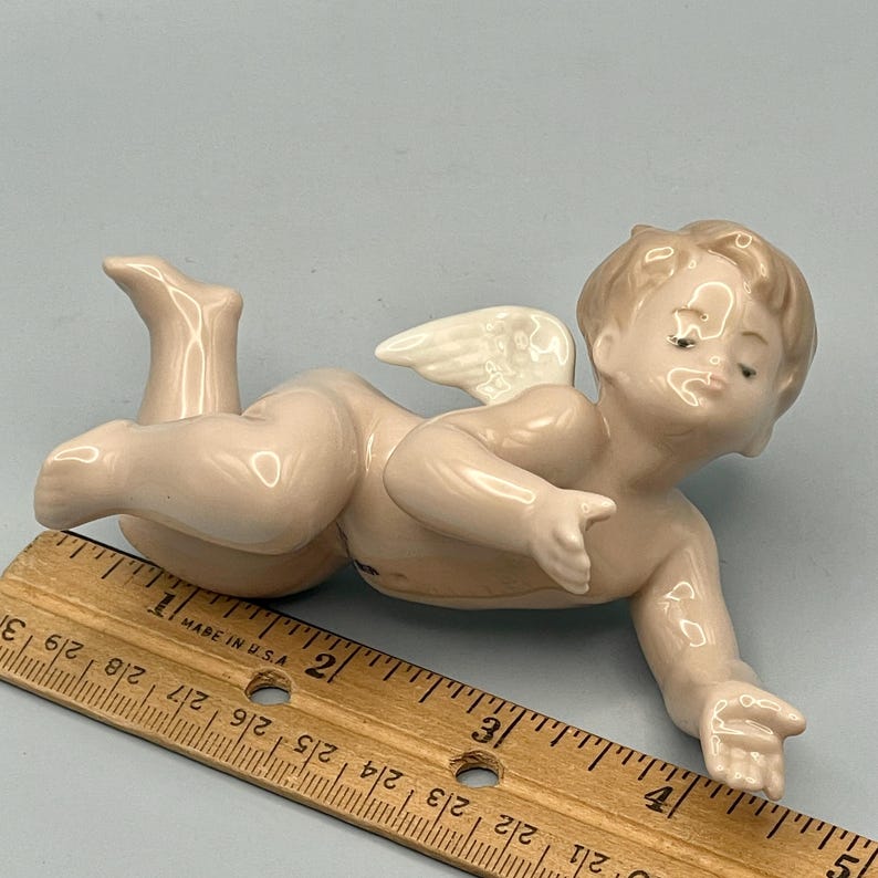 May include: A porcelain figurine of a cherub with closed eyes and small white wings. The cherub is lying down, with one arm extended. The figurine is a light pink color. A wooden ruler is in the foreground.