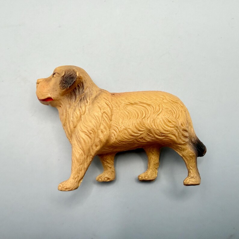 May include: A molded plastic dog figurine in a standing position, featuring a light tan coat with textured fur details, dark brown shading on the ears and tail, and a small red accent on the mouth area, captured from a side profile view.