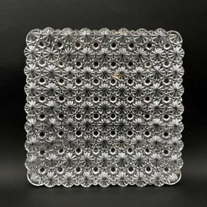 May include: Clear, square glass dish with a detailed, repeating pattern of starbursts and circles. The edges are scalloped. The dish is likely vintage or antique, and is displayed against a black background.