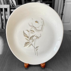 Puede incluir: Una fuente de cerámica blanca con un diseño floral en relieve. La flor es blanca con un centro amarillo, y el tallo y las hojas son marrones y grises. La fuente está sobre un soporte de madera.