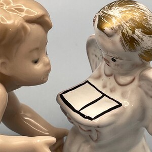 May include: Two porcelain angel figurines. One angel is looking at the other, who is holding a book with a black border. The second angel has gold-colored hair and wings. The figurines are painted in soft, neutral tones.