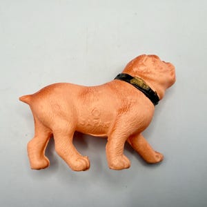 May include: A peach-colored molded plastic dog figurine in a standing position. The dog features a black collar with a gold-colored buckle detail. The word JAPAN is embossed on the side of the dog's body, indicating its origin of manufacture.