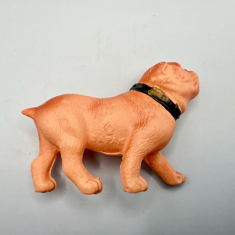 May include: A peach-colored molded plastic dog figurine in a standing position. The dog features a black collar with a gold-colored buckle detail. The word JAPAN is embossed on the side of the dog's body, indicating its origin of manufacture.
