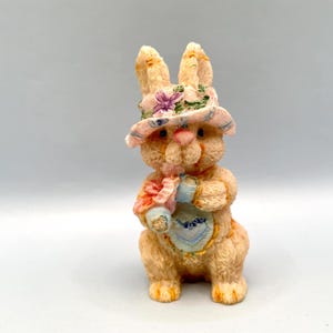 Pode incluir: Estatueta de coelho cor de pêssego com chapéu floral rosa e verde e babete. O coelho segura um ramo de flores e um babete azul e branco com um coração. Decoração de Páscoa.