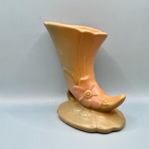 May include: A vintage ceramic vase in the shape of a horn with a pink flower and green leaves. The vase is a light peach color with a darker peach glaze on the horn.