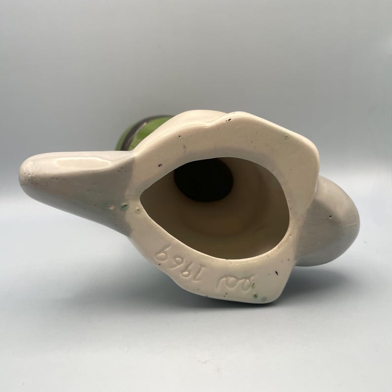 Puede incluir: Una figurita de cer&aacute;mica de un p&aacute;jaro con pintura verde y gris. La figurita est&aacute; de pie sobre una base con las palabras "Roy 1969" escritas en ella.