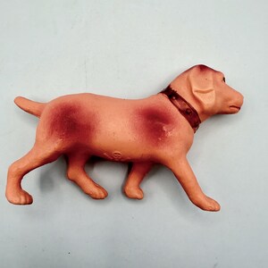 May include: A molded plastic dog figurine in a light peach color with darker reddish-brown shading on the back and sides. The dog is depicted in a standing position, facing right, and features a dark brown collar with small circular details.