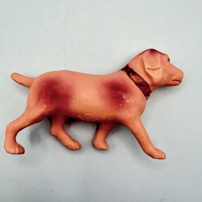 May include: A molded plastic dog figurine in a light peach color with darker reddish-brown shading on the back and sides. The dog is depicted in a standing position, facing right, and features a dark brown collar with small circular details.
