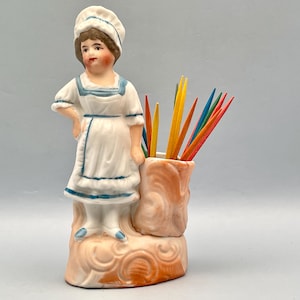 Może przedstawiać: Wazon na wykałaczki w stylu vintage wykonany z porcelany. Przedstawia figurkę kobiety w białym mundurze z niebieskimi wykończeniami. Uchwyt jest w kolorze brzoskwiniowym z teksturowanym wzorem. Uchwyt zawiera kolorowe wykałaczki.