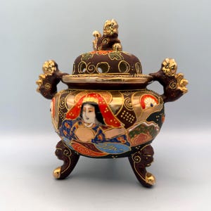May include: A decorative, antique Japanese incense burner. The ceramic vessel features a dark brown lid with gold accents and a detailed painted design. The body of the burner is adorned with a painted woman's portrait and floral motifs. It stands on three ornate legs.