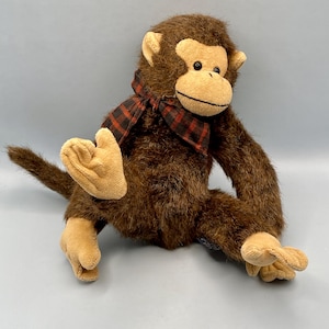 May include: A plush monkey toy with brown fur, tan hands, feet, and face, and a red and black plaid scarf. The monkey is sitting with its legs spread and tail extended. The toy has a friendly expression.