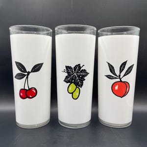 K&ouml;nnte beinhalten: Drei wei&szlig;e, gefrostete Glasbecher mit schwarzen Umrissen von Fruchtmotiven. Der erste Becher hat zwei rote Kirschen mit gr&uuml;nen Stielen und Bl&auml;ttern. Der zweite Becher hat zwei gr&uuml;ne Trauben mit einer schwarzen Rebe und Bl&auml;ttern. Der dritte Becher hat einen roten Pfirsich mit einem gr&uuml;nen Stiel und Bl&auml;ttern.