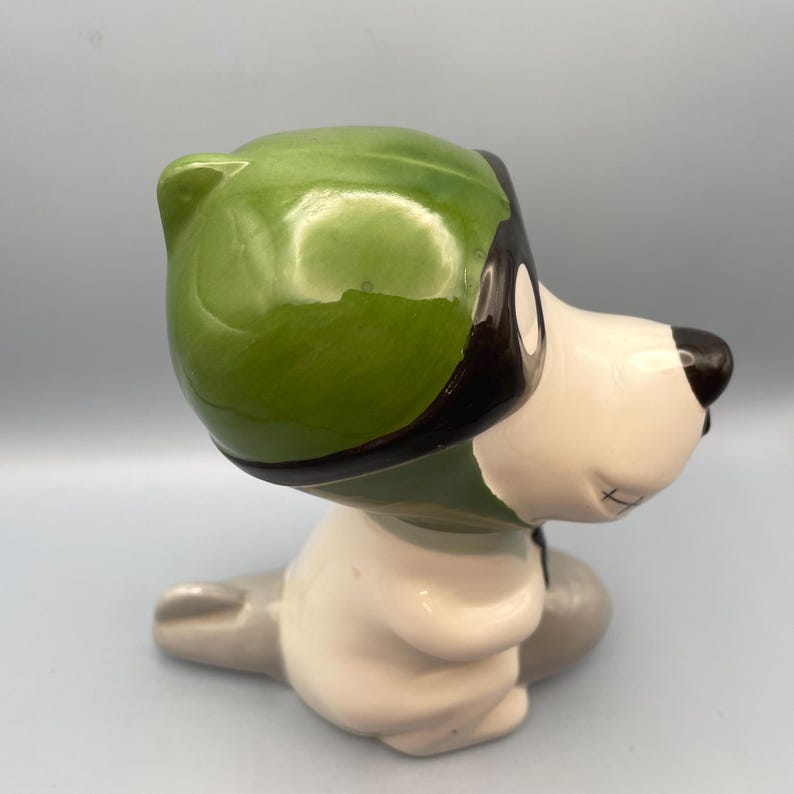 Puede incluir: Una figurita de cer&aacute;mica de un perro con un casco verde y gafas. El perro est&aacute; sentado sobre sus cuartos traseros con la cabeza inclinada hacia un lado.