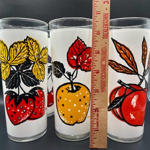 K&ouml;nnte beinhalten: Set aus vier Vintage-Glasbechern mit wei&szlig;em Hintergrund und bunten Fruchtmotiven. Die Motive umfassen Erdbeeren, &Auml;pfel, Kirschen und Pfirsiche.