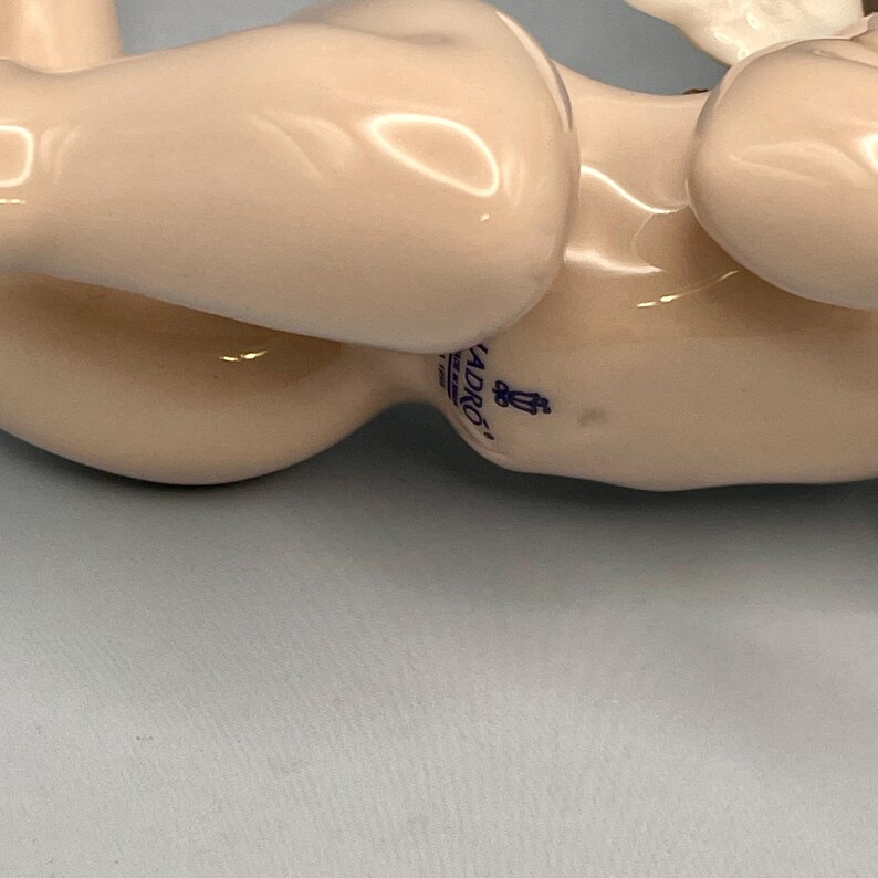 May include: A porcelain figurine in a light beige color. The figurine has a smooth, glossy finish and a curved shape. The word "LADRO" is visible on the figurine, along with a small blue emblem. The figurine is lying on a light blue surface.