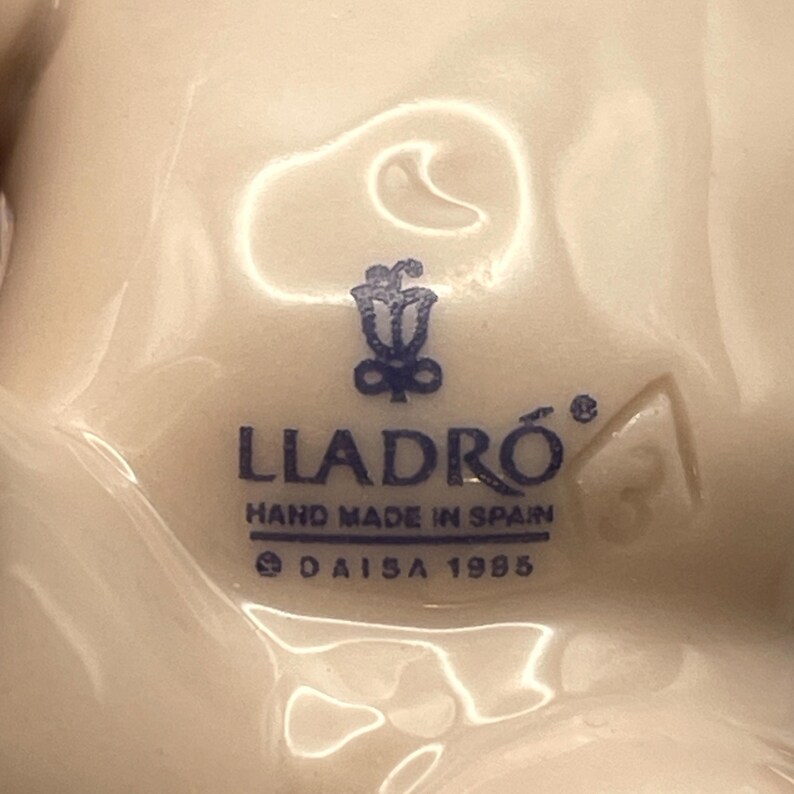 May include: Close-up of a white porcelain figurine base with the blue text "LLADR&Oacute; HAND MADE IN SPAIN &copy; DAISA 1985". The logo features a floral design. A number "3" is visible.