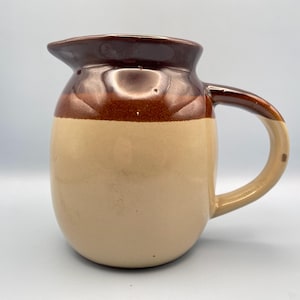Può includere: Una brocca in ceramica con bordo e manico marroni e corpo beige. La brocca ha una forma arrotondata e un beccuccio per versare. Il manico è curvo e attaccato al lato.