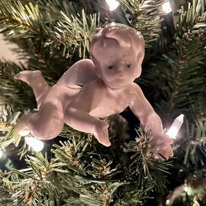 May include: A vintage Christmas tree ornament in the shape of a crawling baby. The ornament is made of porcelain and is a pale pink color. The baby is reaching out with one hand. The ornament is hanging on a green Christmas tree with white lights.
