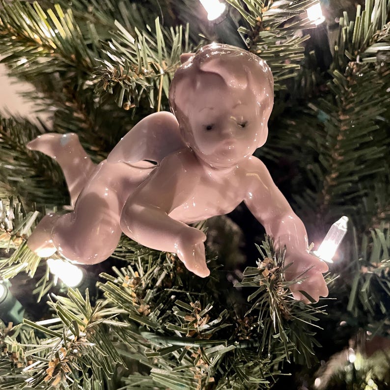 May include: A vintage Christmas tree ornament in the shape of a crawling baby. The ornament is made of porcelain and is a pale pink color. The baby is reaching out with one hand. The ornament is hanging on a green Christmas tree with white lights.