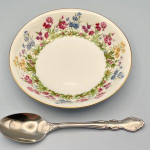 May include: A cream-colored bowl with a floral design in pink, blue, and yellow, and a gold rim. A silver spoon with a decorative handle is next to the bowl. The bowl is likely for serving soup or cereal.