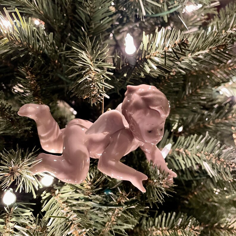 May include: A pink ceramic cherub Christmas ornament with outstretched arms and small wings. The cherub is attached to a gold string and is hanging on a green Christmas tree with white lights.