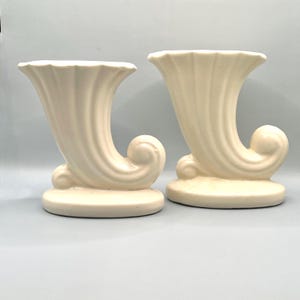 Può includere: Due vasi in ceramica color crema a forma di cornucopia. Ogni vaso ha una parte superiore svasata, un corpo curvo e una base arrotondata. I vasi sono identici per forma e dimensioni e sono posti uno accanto all'altro.