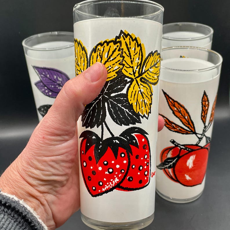 Four vintage drinking glasses with a white background and colorful fruit and leaf designs. The glasses feature strawberries, leaves, and a yellow flower design.