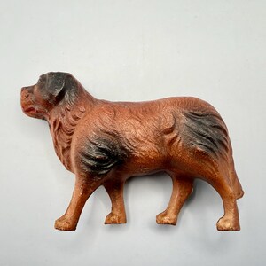 May include: A cast metal figurine of a dog standing in profile. The dog has a textured brown coat with dark black shading on the ears, muzzle, and hindquarters. The figure is depicted in a standing position with four legs visible and a short tail.