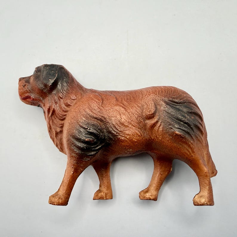 May include: A cast metal figurine of a dog standing in profile. The dog has a textured brown coat with dark black shading on the ears, muzzle, and hindquarters. The figure is depicted in a standing position with four legs visible and a short tail.