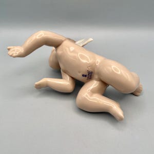 May include: A porcelain figurine of a crawling cherub with a small white wing. The cherub is in a crawling position, with its arms and legs extended. The figurine is a light flesh tone with a glossy finish. The base of the figurine has a blue stamp.