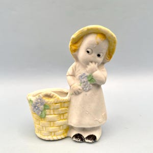 May include: A vintage ceramic figurine of a young child in a dress and hat, with a yellow basket. The child is holding a small bouquet of flowers. The basket is yellow with blue and green floral accents. The figurine is painted in soft, pastel colors.