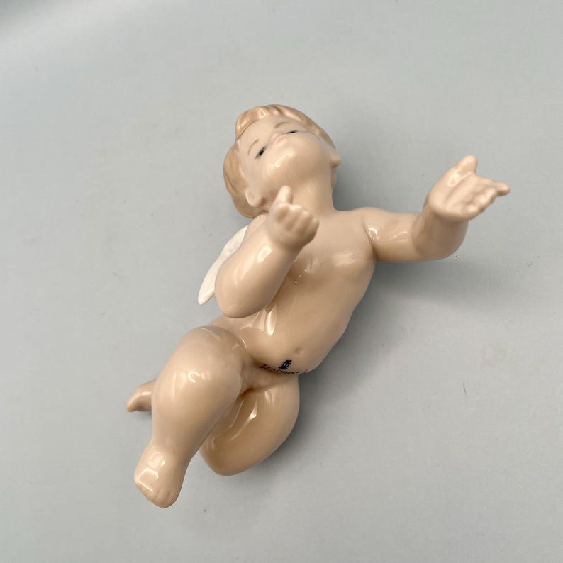 May include: A porcelain figurine of a cherub with blonde hair and small white wings. The cherub is in a reclining pose with arms outstretched, and is painted in a flesh tone with subtle shading. The figurine is on a light blue surface.