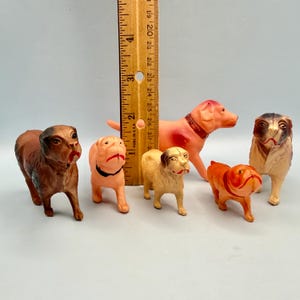May include: A collection of six small dog figurines in various colors including brown, tan, and orange, positioned next to a wooden ruler that shows the tallest figure stands at approximately 3 inches in height. Each dog has painted features.