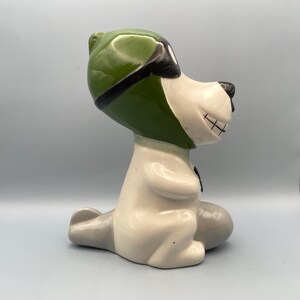 A ceramic figurine of a dog wearing a green aviator hat and goggles. The dog is sitting on a gray plane and has a white body with black eyes and a black mouth.