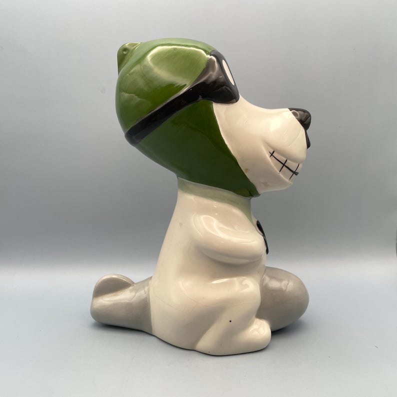 A ceramic figurine of a dog wearing a green aviator hat and goggles. The dog is sitting on a gray plane and has a white body with black eyes and a black mouth.