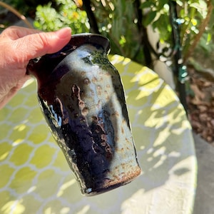 May include: A ceramic tumbler featuring a glossy glaze with a blend of deep navy blue, charcoal, and earthy brown tones. The surface has a textured, dimpled appearance with a smooth, reflective finish and a slightly flared rim.