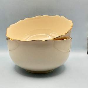 May include: Two stacked cream-colored bowls with a smooth, glossy finish and scalloped edges. Each bowl features a thin, metallic gold trim along the rim, highlighting the curved, wavy design of the upper edge of the ceramic kitchenware.