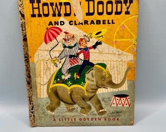 Puede incluir: Libro infantil vintage titulado "Howdy Doody and Clarabell". La portada presenta una escena de circo con un payaso, un niño y un elefante. El libro tiene un fondo amarillo con texto azul y rojo.