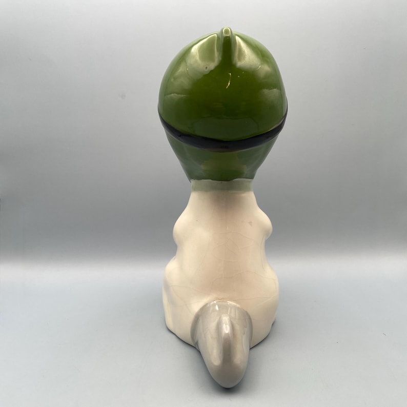 Puede incluir: Una figurita de cer&aacute;mica de un animal sentado con un sombrero verde. El animal tiene un cuerpo blanco y una cola gris.