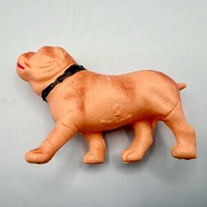 May include: A light tan molded plastic dog figurine in a standing position. The dog has a black collar around its neck and a small red painted detail on its mouth. The surface of the figurine features textured fur details and a small embossed mark.