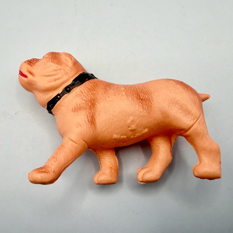 May include: A light tan molded plastic dog figurine in a standing position. The dog has a black collar around its neck and a small red painted detail on its mouth. The surface of the figurine features textured fur details and a small embossed mark.