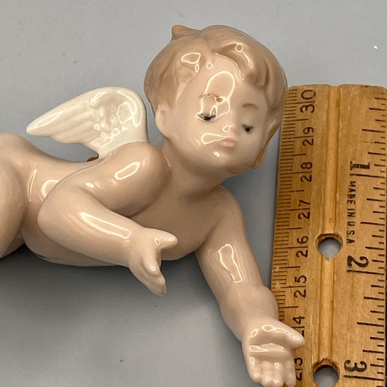 May include: A porcelain cherub figurine with a light skin tone, small wings, and a serene expression. The cherub is lying down, with one arm extended. A wooden ruler, marked in inches, is next to the figurine.