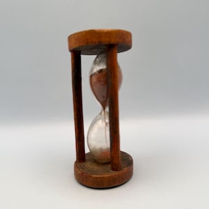 Op de afbeelding: Een houten zandloper met een bruin frame en basis, met bruin zand in een heldere glazen bol. De zandloper is een klassiek tijdmeetapparaat, met een eenvoudig, elegant ontwerp.