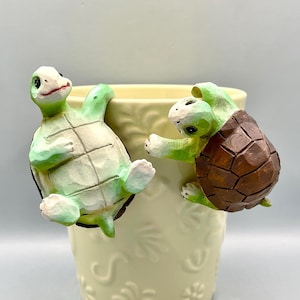 May include: Two decorative turtle figurines, one with a brown shell and the other with a green shell, are attached to the rim of a cream-colored ceramic pot. The turtles appear to be climbing the pot, adding a whimsical touch.